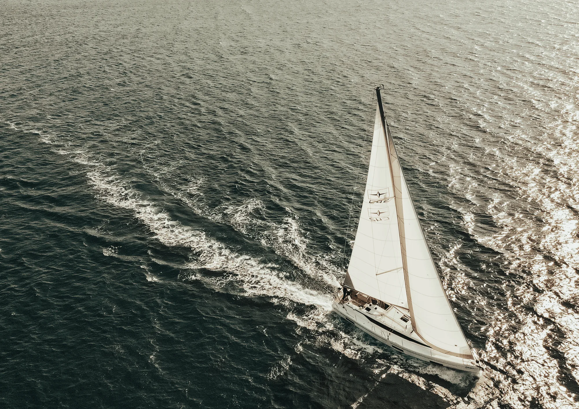 A white sailboat seen from above sailing across calm blue water under the sunlight, leaving a white wake behind.