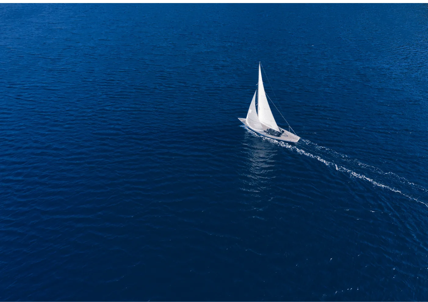 Aerial view of a sailboat sailing alone across the deep blue Atlantic Ocean, leaving a white wake behind.