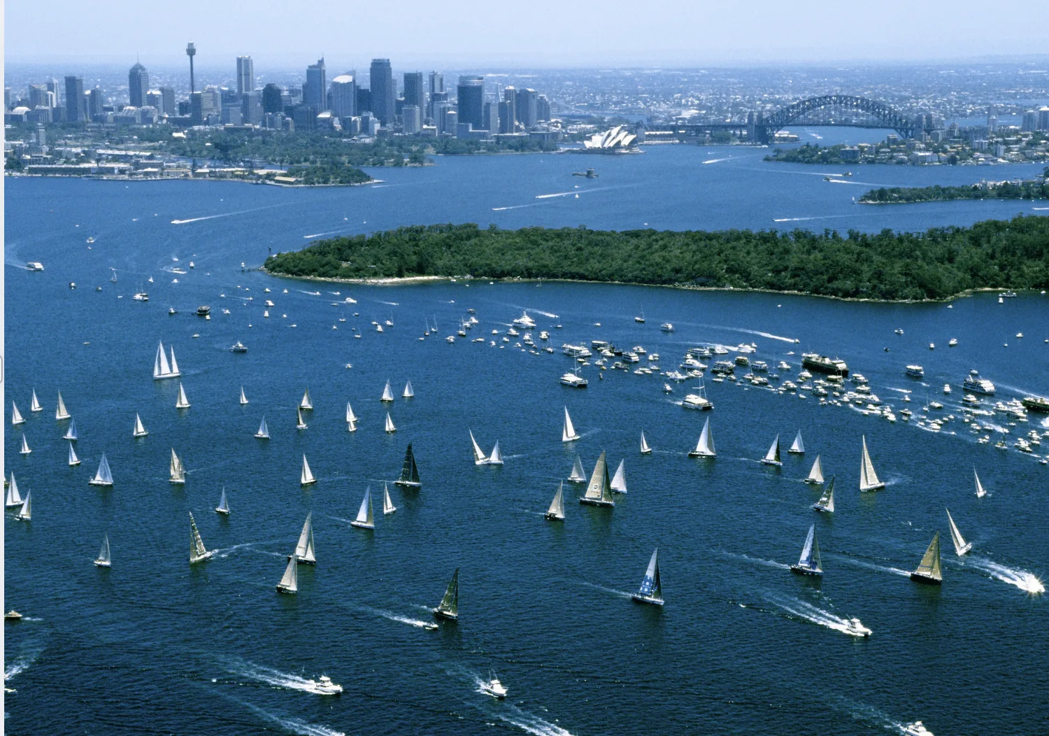 Aerial view of the Sydney Hobart Yacht Race start in Sydney Harbour with dozens of sailboats heading toward the open ocean and the Sydney skyline in the background.